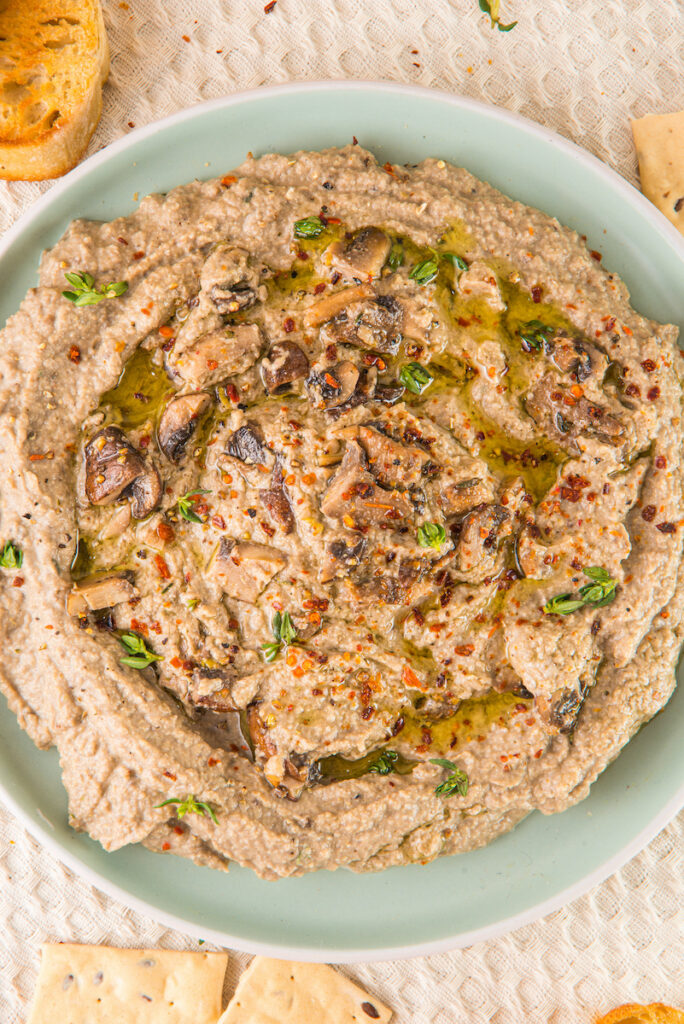 A plate of creamy mushroom hummus, elegantly garnished with a drizzle of oil, fresh herbs, and a sprinkle of chili flakes, is beautifully surrounded by rustic bread slices and crispy crackers.