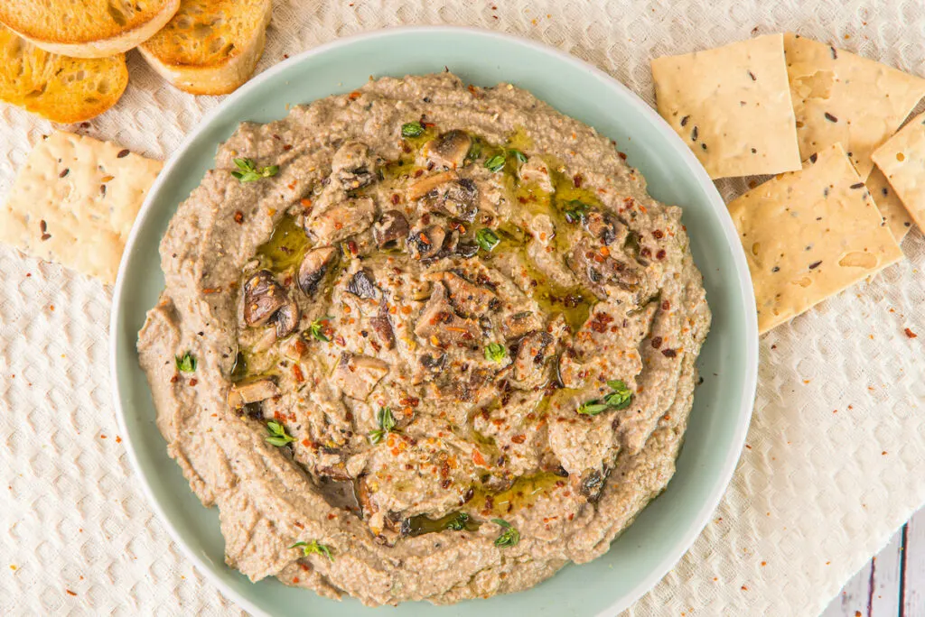 A plate of eggplant dip garnished with herbs and spices, accompanied by toasted bread and crackers, sits beside a bowl of earthy mushroom hummus.