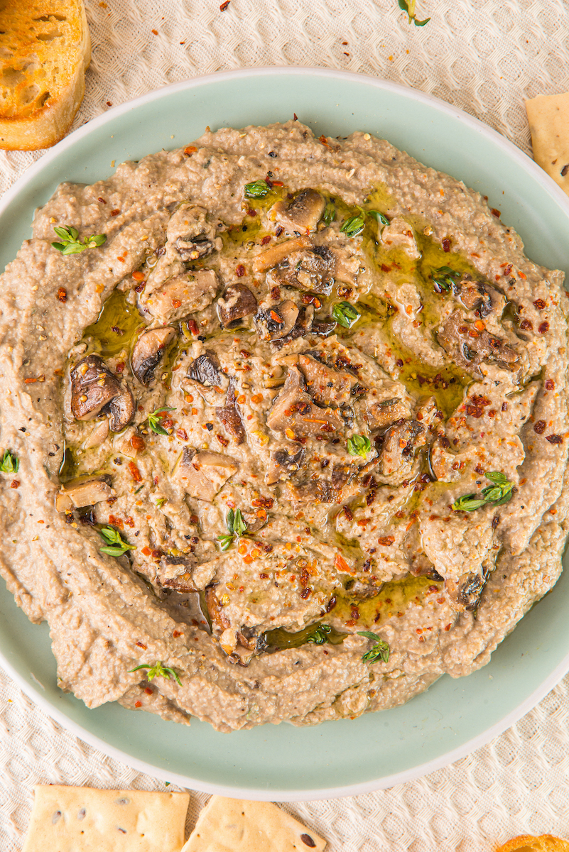 A plate of creamy mushroom hummus, elegantly garnished with a drizzle of oil, fresh herbs, and a sprinkle of chili flakes, is beautifully surrounded by rustic bread slices and crispy crackers.