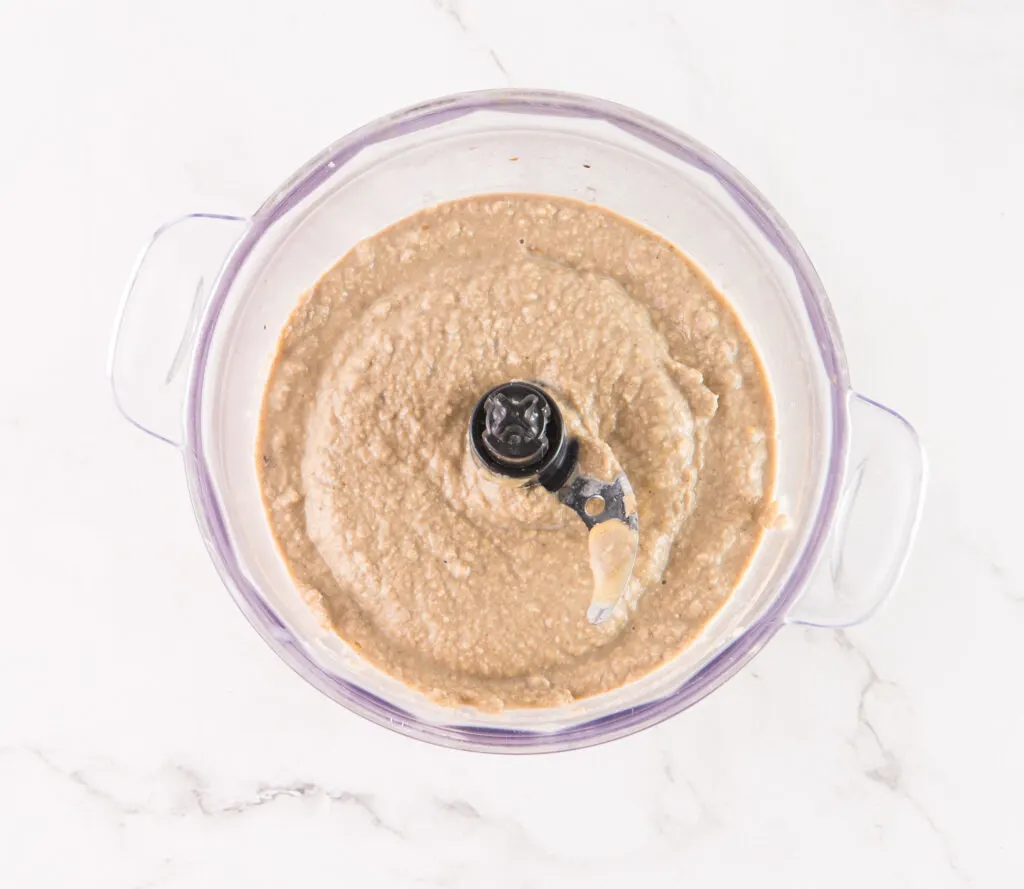 A glass food processor bowl filled with a creamy, beige mushroom hummus sits elegantly on a marble surface.