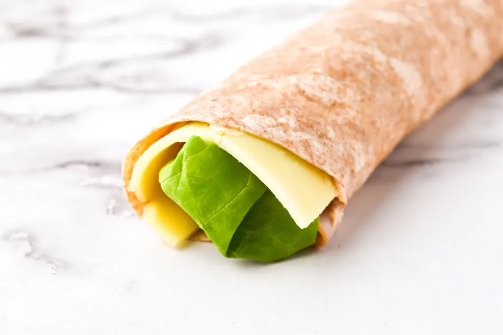 A whole wheat wrap with cheese, lettuce, and tender turkey pinwheel slices rests elegantly on a marble surface.