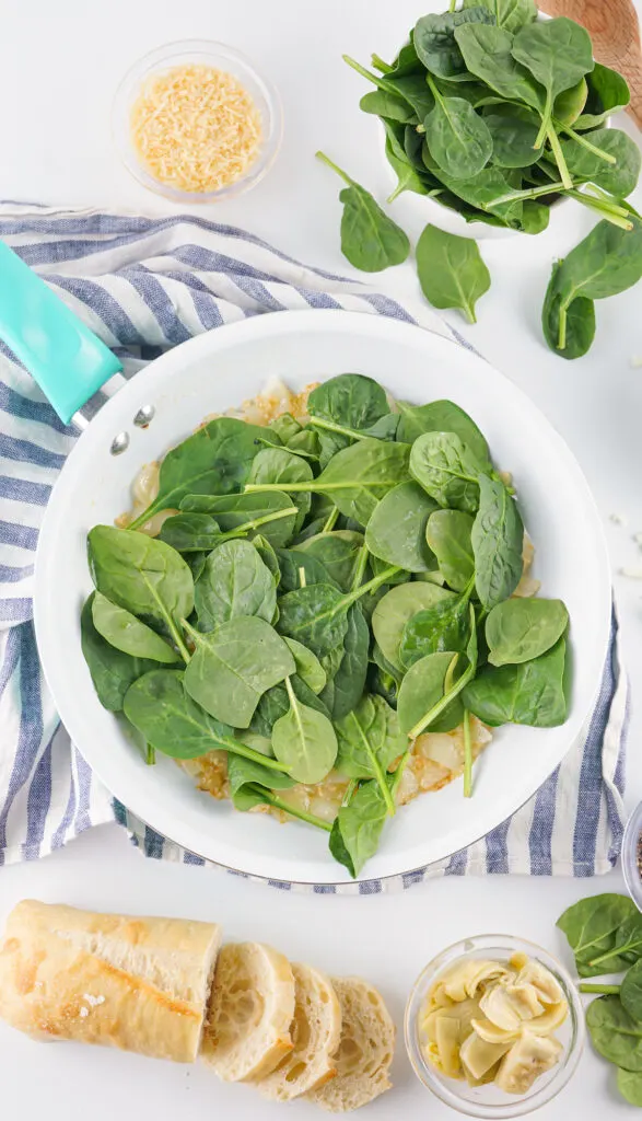 A skillet filled with fresh spinach on a striped towel, surrounded by grated cheese, sliced baguette, and a bowl of artichoke hearts.