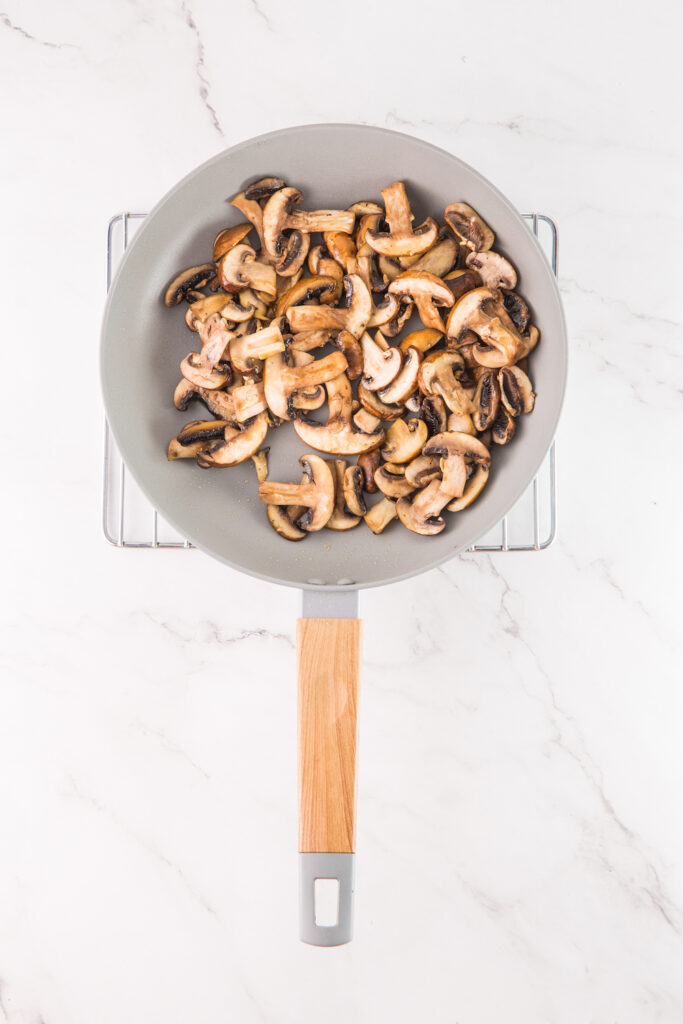 Sliced mushrooms sautéed in a frying pan with a wooden handle rest on a marble surface, hinting at their future transformation into creamy mushroom hummus.