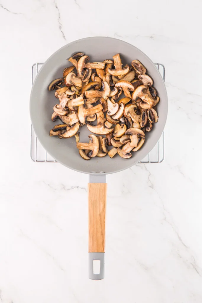 Sliced mushrooms sautéed in a frying pan with a wooden handle rest on a marble surface, hinting at their future transformation into creamy mushroom hummus.