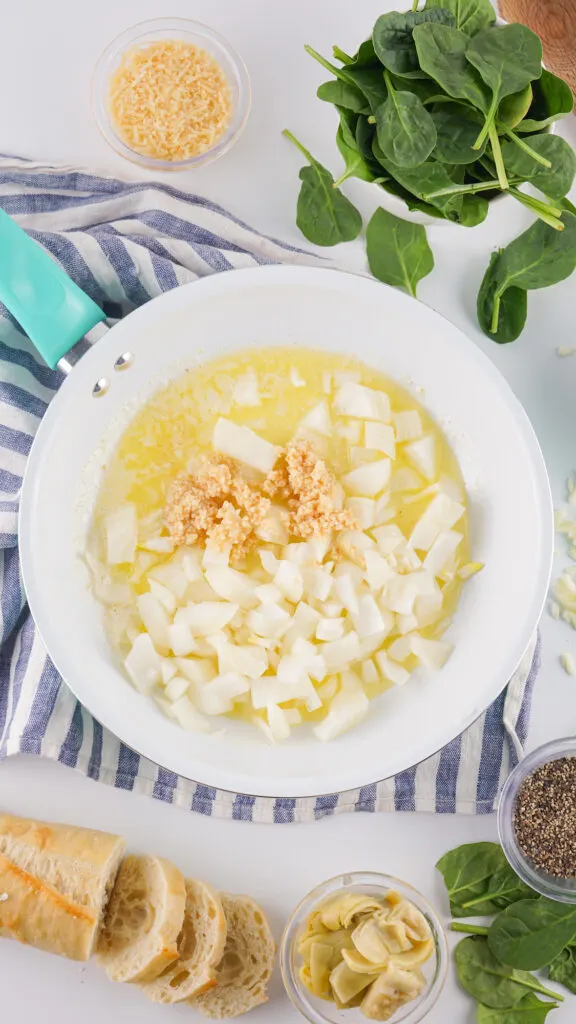 A frying pan with diced onions and garlic in butter. Nearby are fresh spinach, grated cheese, artichoke hearts, black pepper, and slices of baguette.