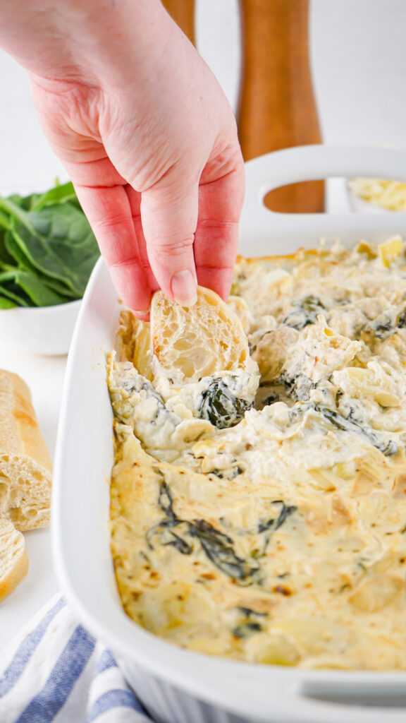 A hand dipping bread into a creamy spinach and artichoke dip in a white dish.