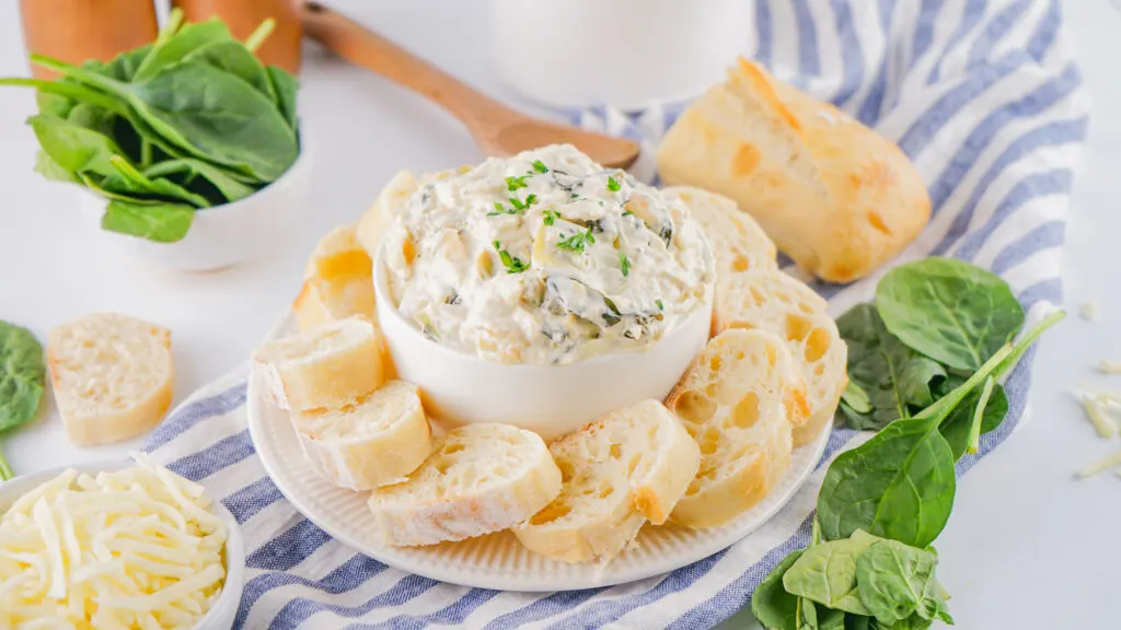 A bowl of creamy dip, garnished with herbs, surrounded by slices of bread on a striped cloth. Fresh spinach leaves and shredded cheese are beside the plate.