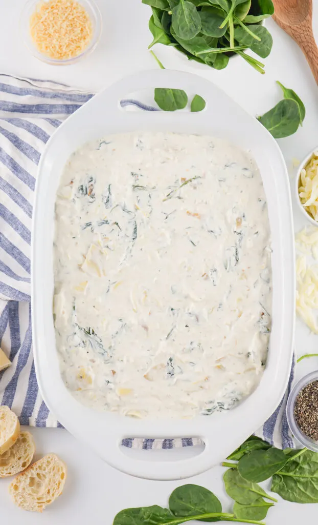 A white dish filled with a creamy spinach and artichoke mixture. Surrounded by fresh spinach, bread slices, grated cheese, and a striped cloth.