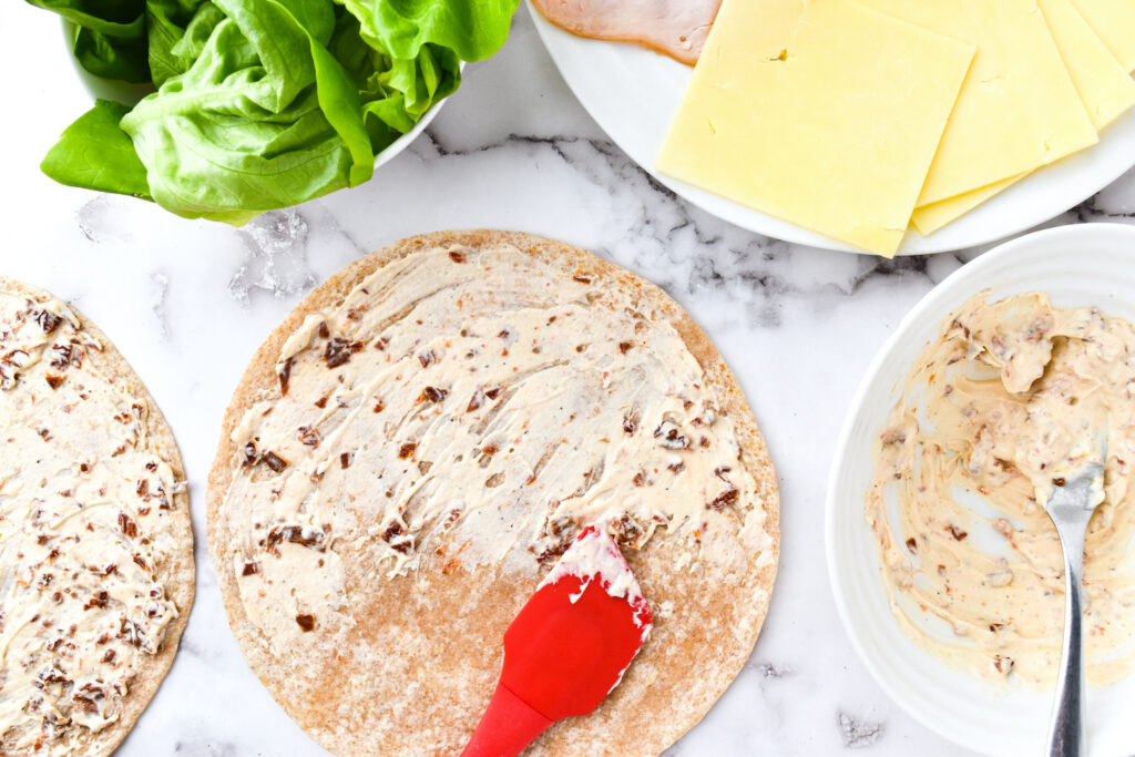 Tortilla being spread with a creamy mixture next to lettuce, cheese slices, and a plate with more spread, ready to be rolled into delicious turkey pinwheels.