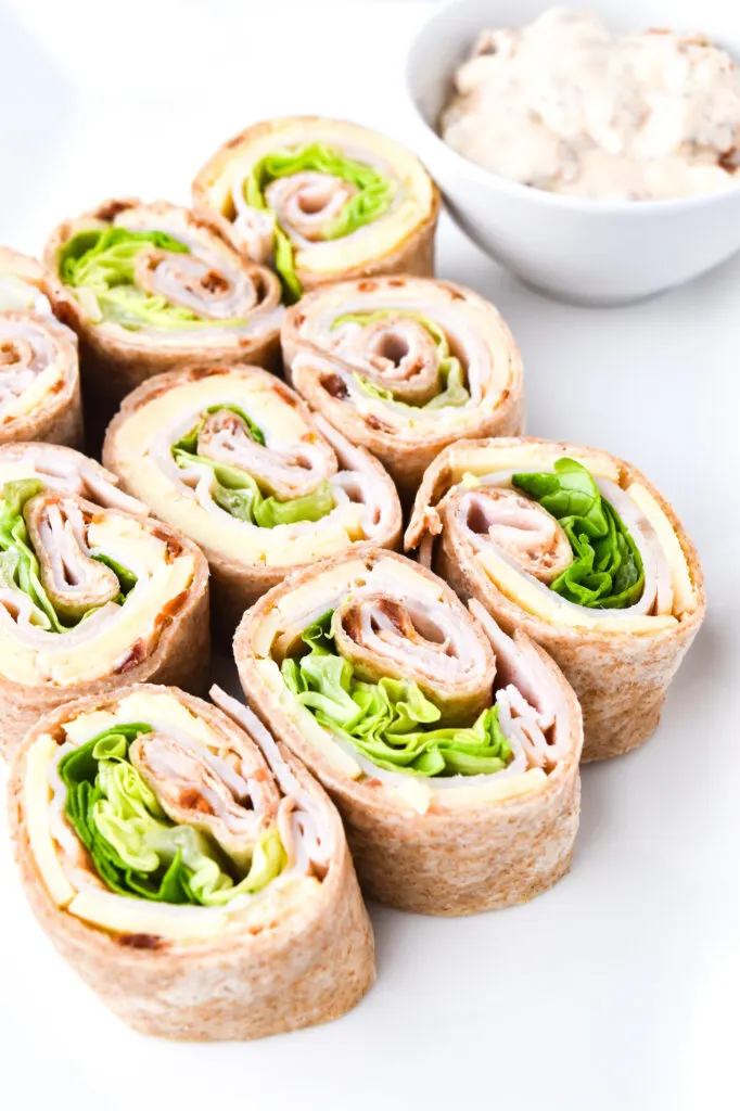 Pinwheel sandwiches filled with turkey, lettuce, and cheese are arranged on a plate. A small bowl of dip is beside them.